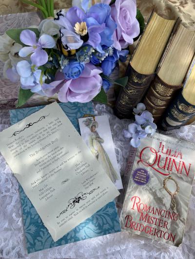 Overhead view of a tabletop with a vase of purple flowers, three old books, a paperback version of Julia Quinn's Romancing Mister Bridgerton, an invitation to a Bridgerton Ball, and a bookmark featuring a woman dressed in period fashion