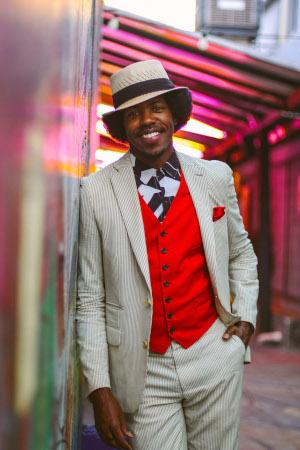 A Black man wearing a tan suit with red vest and a tan brimmed hat leans against a wall with his hand in his pocket, smiling at the camera