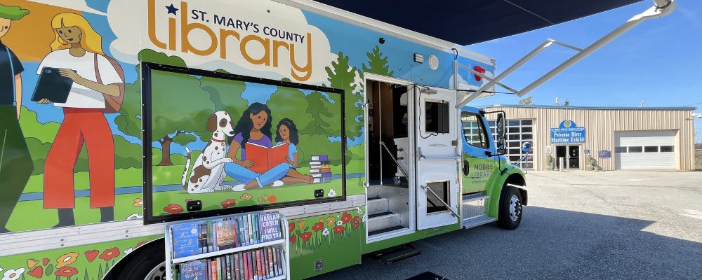 The Mobile Library, with Library logo and artwork featuring two people standing and talking and a woman reading to a dog and child, parked outside the Potomac River Maritime Exhibit building. A book cart of hardcover books sits next to the vehicle.