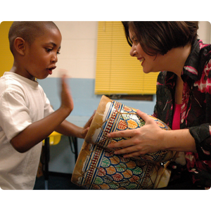 A woman with short brown hair holds a drum while a young Black child hits the drum