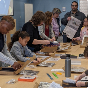 A group of children and adults stand around a table making eclipse viewers using cardboard, newspaper, and aluminum foil.