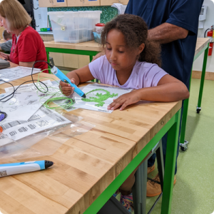 A young girl in a purple shirt uses a 3D pen to create a green lizard