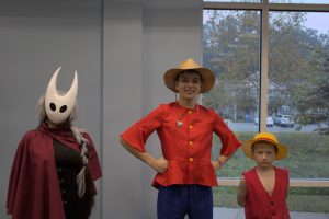 Three young cosplayers, one in a Hollow Knight mask, one in a red shirt, blue pants, and tan brimmed hat, and one in a tan brimmed hat and red vest.