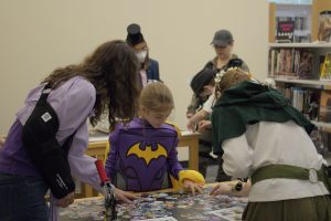 Several people standing over a table with cutouts for pin buttons and a button maker. A young girl is dressed in a purple batman suit.