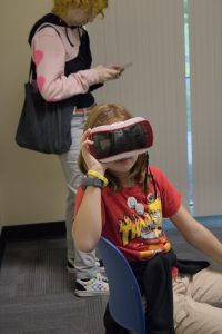 A young girl sits in a chair holding a virtual reality viewer to her face