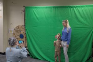 A blonde woman wearing a blue shirt and grey pants stands with a young girl in an animal print outfit holding a face mask in front of a green screen while another woman takes their photo