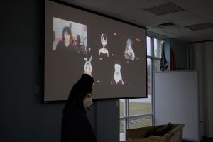 A projection screen in a dark room shows five figures - one young man and four female anime-style avatars