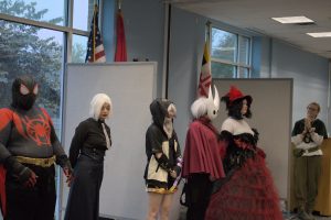 A group of cosplayers, including Spiderman, Hollow Knight, a character in black with white hair, and a woman in a red and black dress.