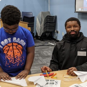 A man in a black jacket and a young boy in a blue and orange shirt make paper airplanes