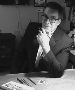 A black and white photo of a man wearing glasses and a dark sports coat, sitting at a table looking at the camera