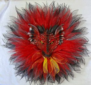 A red dragon with a wreath around its face made of red and black feathers. The face is painted red and black and the eyes are glowing yellow.