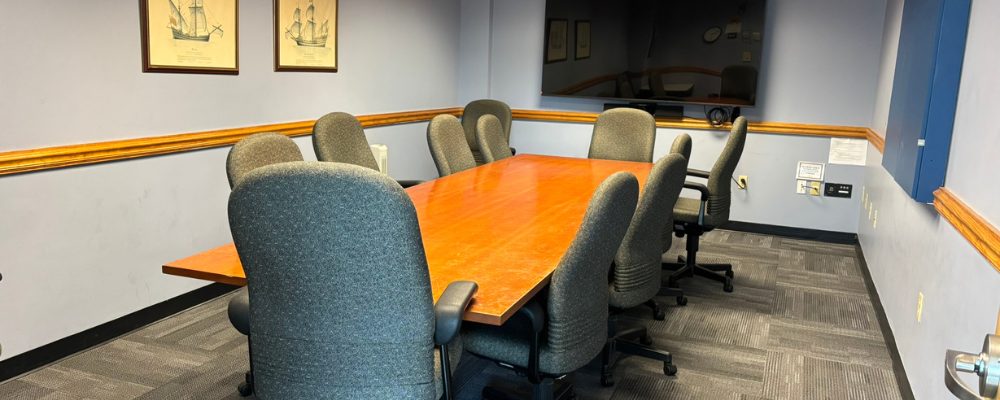 A conference table with 10-12 office chairs. A wall mounted tv is at the front of the room.