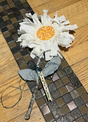 A daisy with petals made from torn muslin strips around a yellow patterned center, a stem made from a stick, and leaves cut from black and white houndstooth fabric on a background of light colored natural wood and dark brown and black small tiles