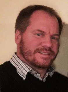 A man with short brown hair and a beard and mustache wearing a collared shirt under a sweater looks at the camera