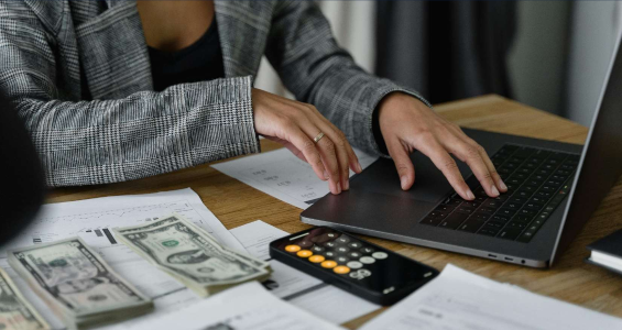 A woman in a black and white jacket sits at a table with a laptop, calculator, papers, and two stacks of money