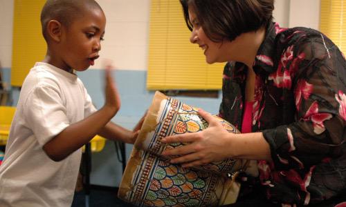 A woman with short brown hair holds a drum and a young Black child hits the drum