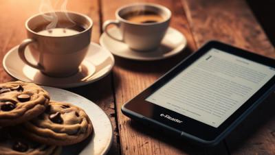 An ereader sits on a table next to two cups of coffee and a plate of cookies