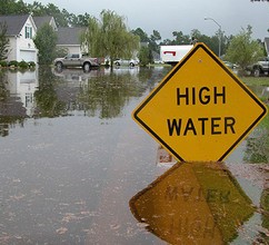 A yellow High Water sign next to a mailbox in a flooded neighborhood with houses and cars in the background