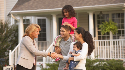 A blonde haired woman in a tan blazer shakes hands with a woman with long dark hair holding a baby outside a house. Next to the woman with dark hair is a smiling man with a young girl on his shoulders.