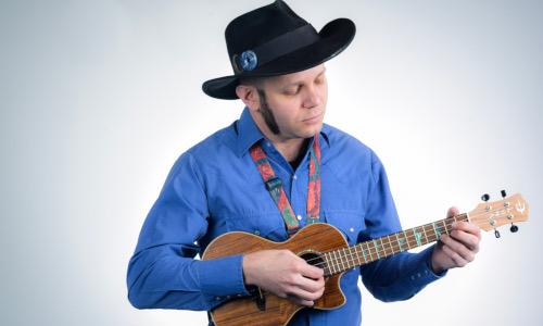 A man in a blue shirt and a cowboy hat strums a ukulele