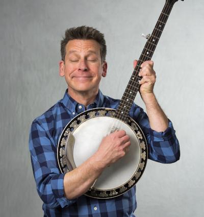 A man in a blue and black plaid shirt stands with his eyes closed, strumming a banjo