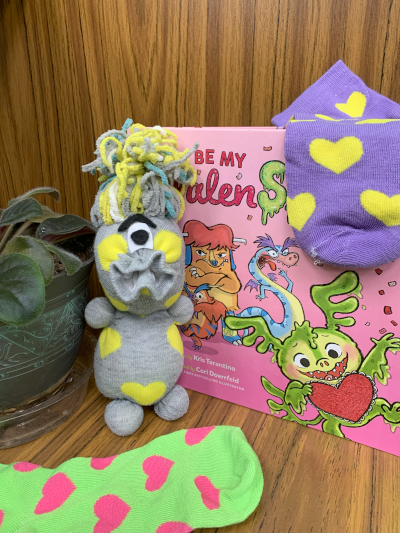 A love monster made out of a sock, pom poms, and other craft materials sites next to a picture book about Valentine's Day