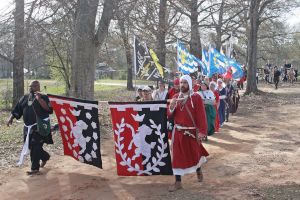 A parade of people in medieval dress walking through trees waving colorful flags and banners