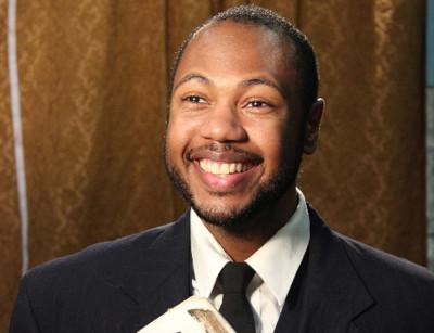 A Black man with facial hair wearing a suit jacket and tie stands smiling in front of a gold curtain