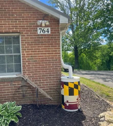 A brick house with a rain barrel with the Maryland flag painted on it attached to one of the gutters