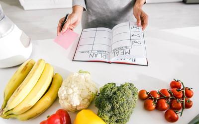 A countertop covered in fruits and vegetables and a meal planning book. A woman holding a piece of paper and a pen stands over the book.