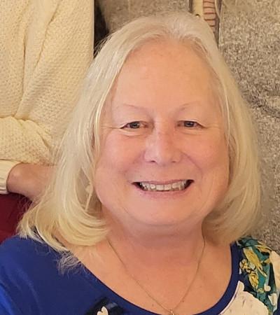 Headshot of Susan Youhn, a woman with shoulder length white hair, wearing a blue shirt and smiling