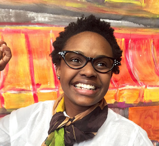 A Black woman with short hair and glasses stands smiling in front of an orange and yellow background